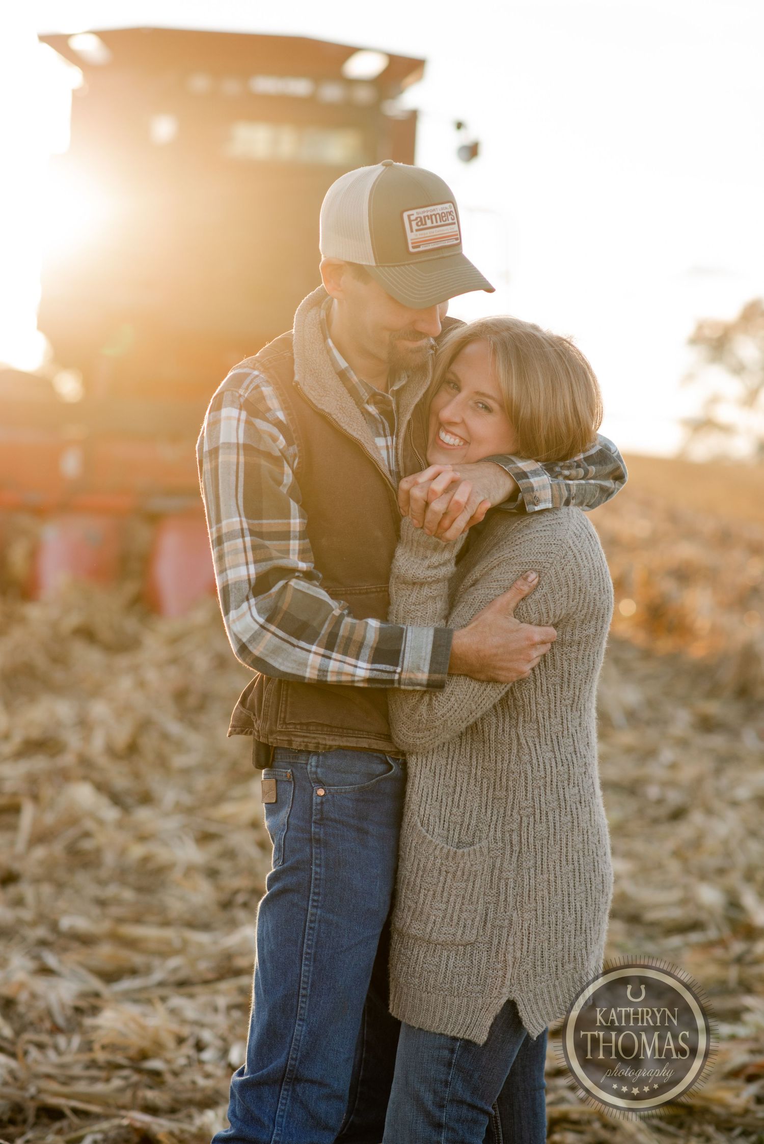 A loving couple embraces in front of farm equipment during golden hour sunset.