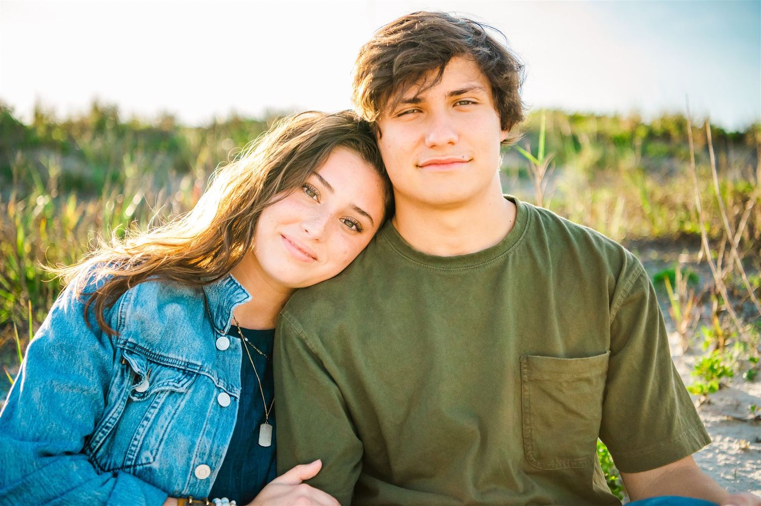 A young couple sits together in a sunlit field during golden hour wearing denim jacket and olive t-shirt.