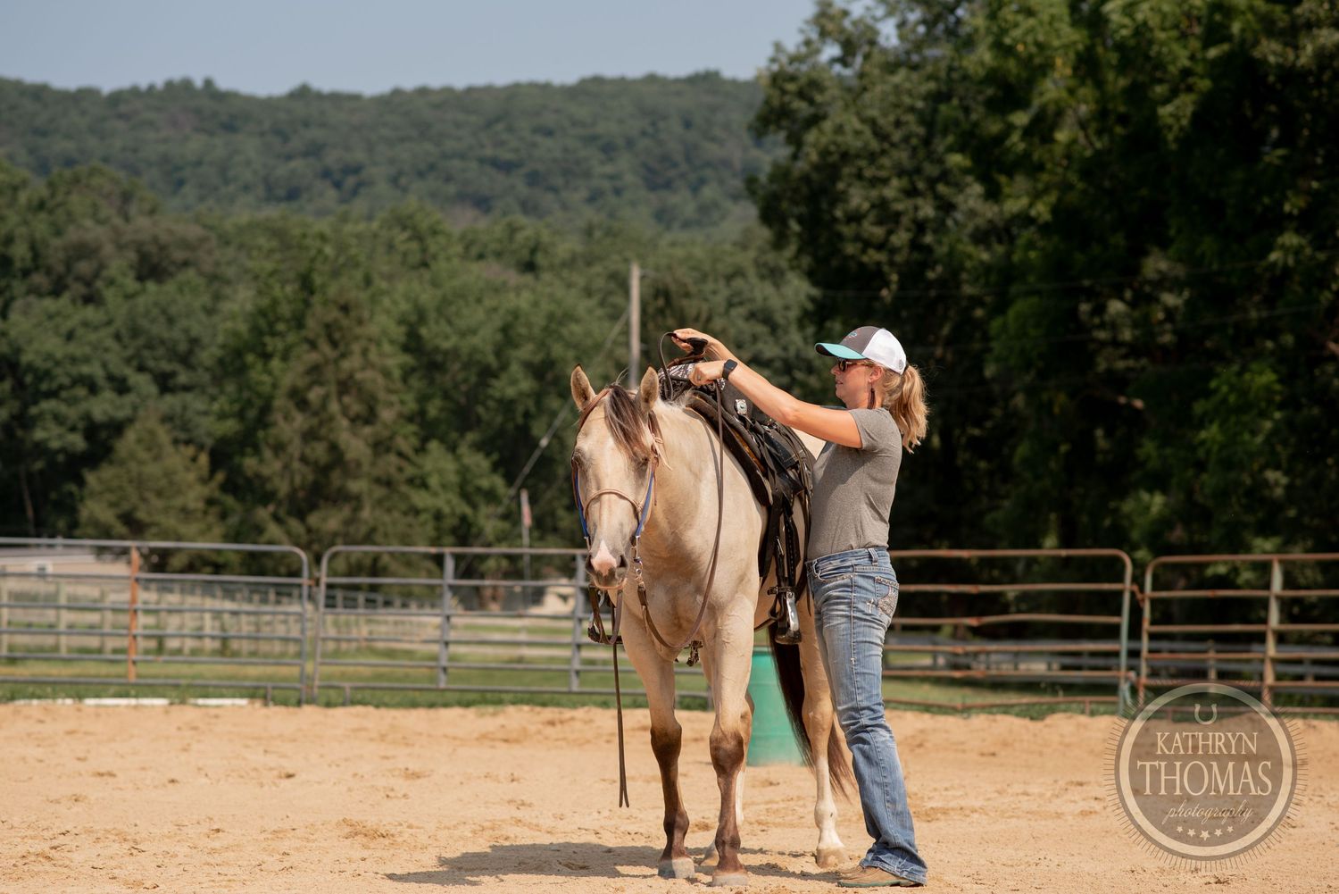 Horse being groomed in outdoor arena with wooded hills in background.