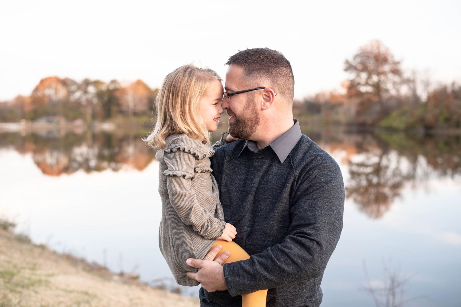 Dad and little girl give each other a nose kiss in front of a lake in this photograph.