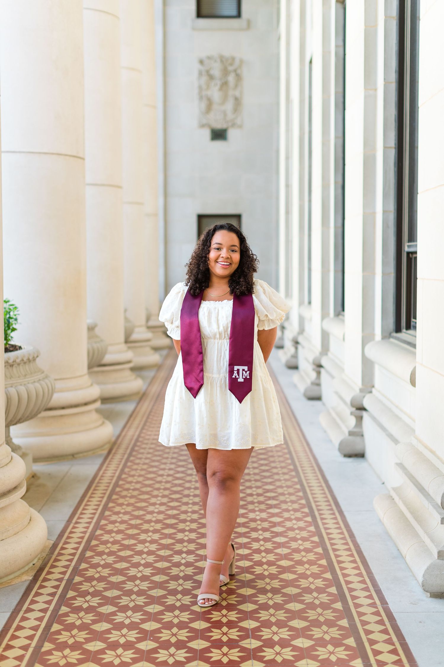 Graduate in white dress and maroon stole walks along decorative tiled colonnade.