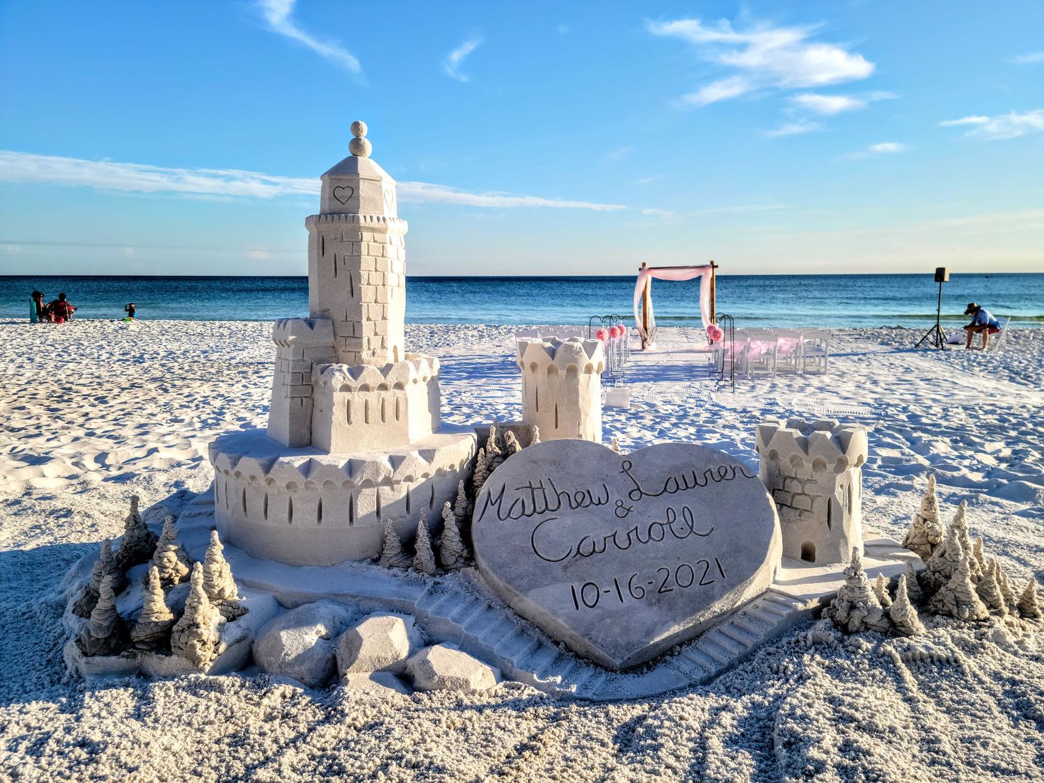 A detailed sandcastle with lighthouse and heart shapes carved into it on a white sandy beach.