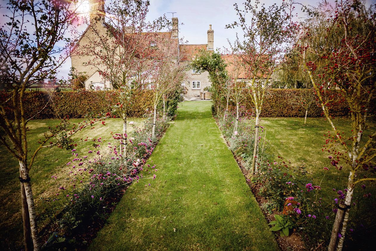 Manicured English garden path lined with young trees leading to a traditional stone cottage.