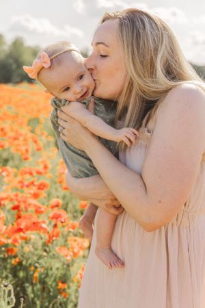 Blonde mother in pink dress kissing baby girl in green outfit in a blooming poppy field.