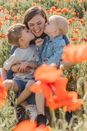 Mother laughing joyfully as two young boys kiss her cheeks in a vibrant red poppy field.