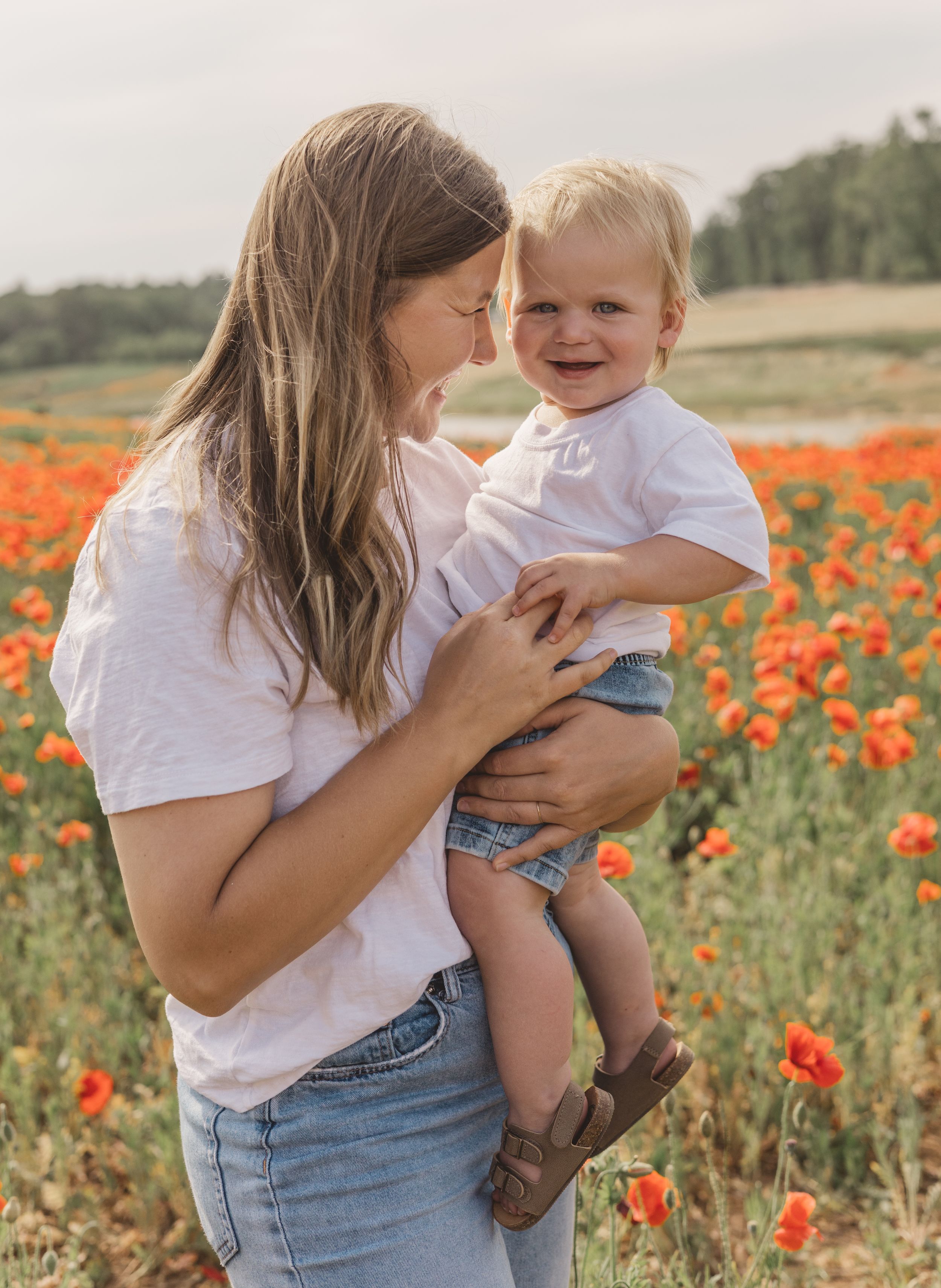 Mother holding smiling toddler in a vibrant red poppy flower field on a sunny day.