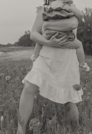 Mother in white dress carrying toddler through poppy field, tender black and white family photography moment.