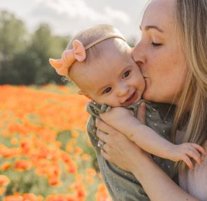 Loving mother kissing adorable baby girl with pink bow headband in an orange poppy field.