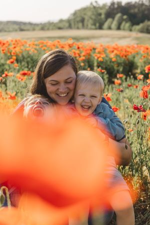 Happy mother hugging laughing toddler boy in a stunning red poppy flower field outdoors.