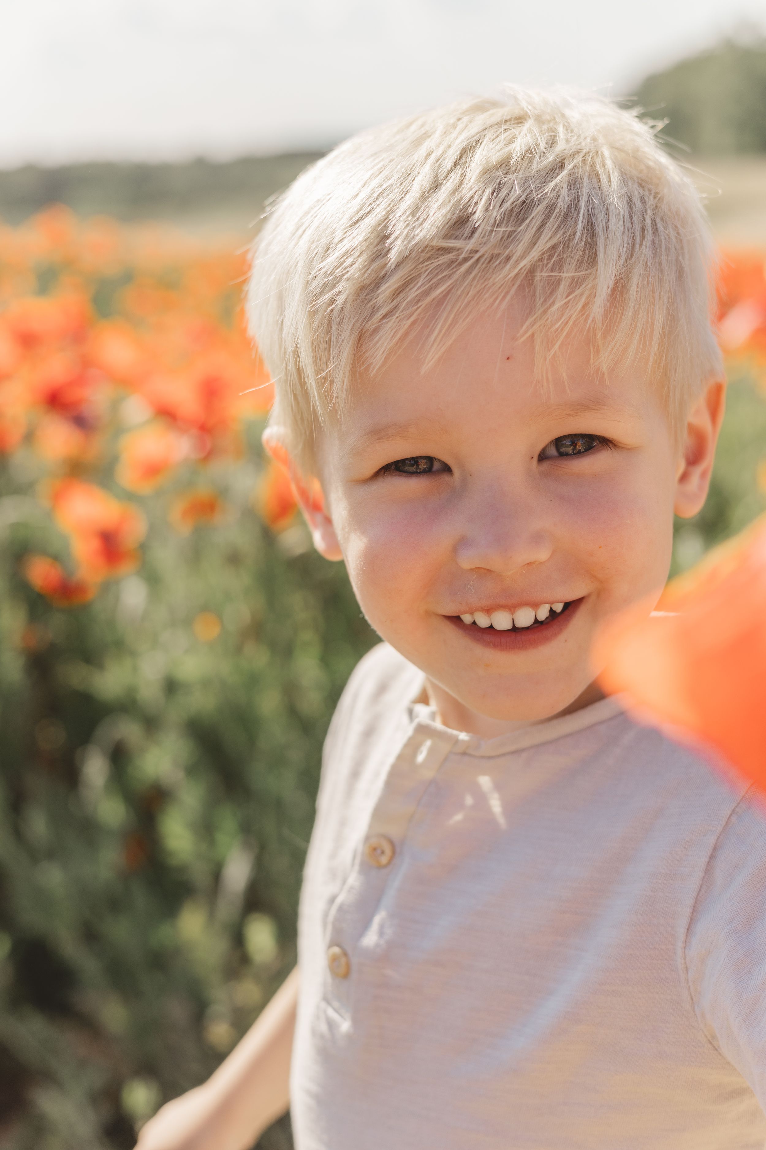 Adorable smiling blonde toddler boy in white shirt standing in vibrant orange poppy flower field outdoors.