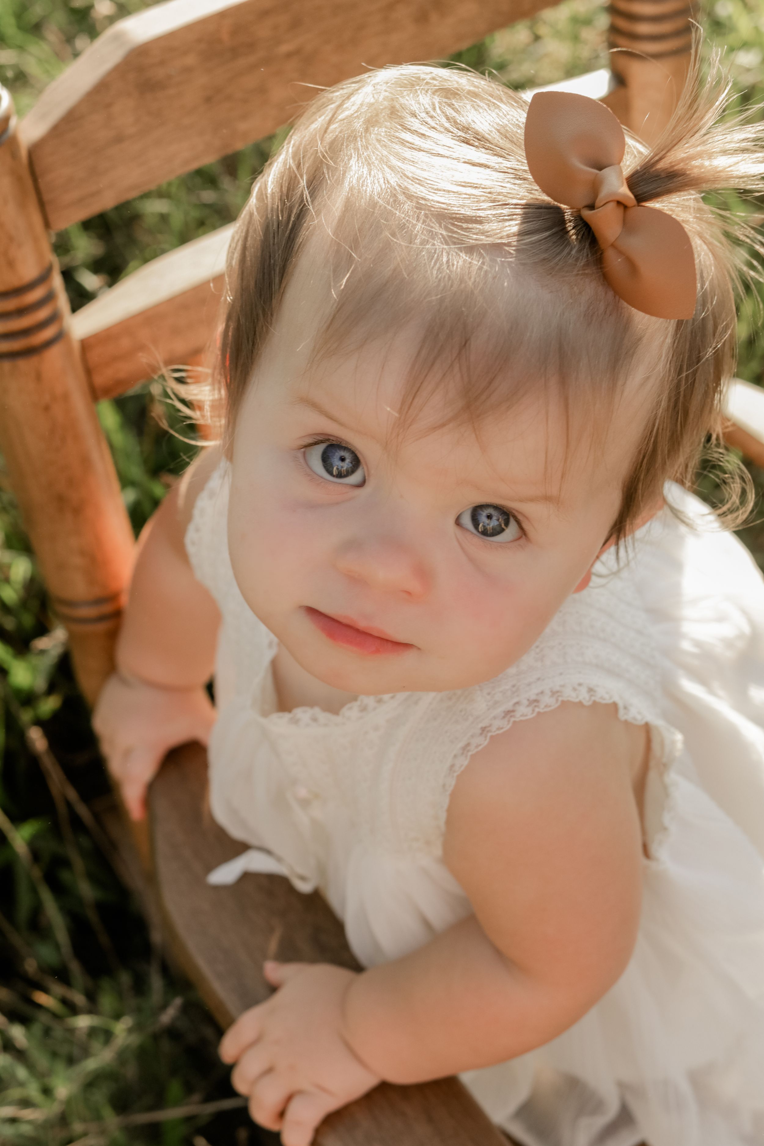 Adorable baby girl with blue eyes in white dress and bow sitting in wooden chair outdoors.