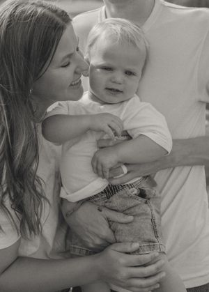 Mother smiles at baby boy held in parents arms in black and white family portrait.