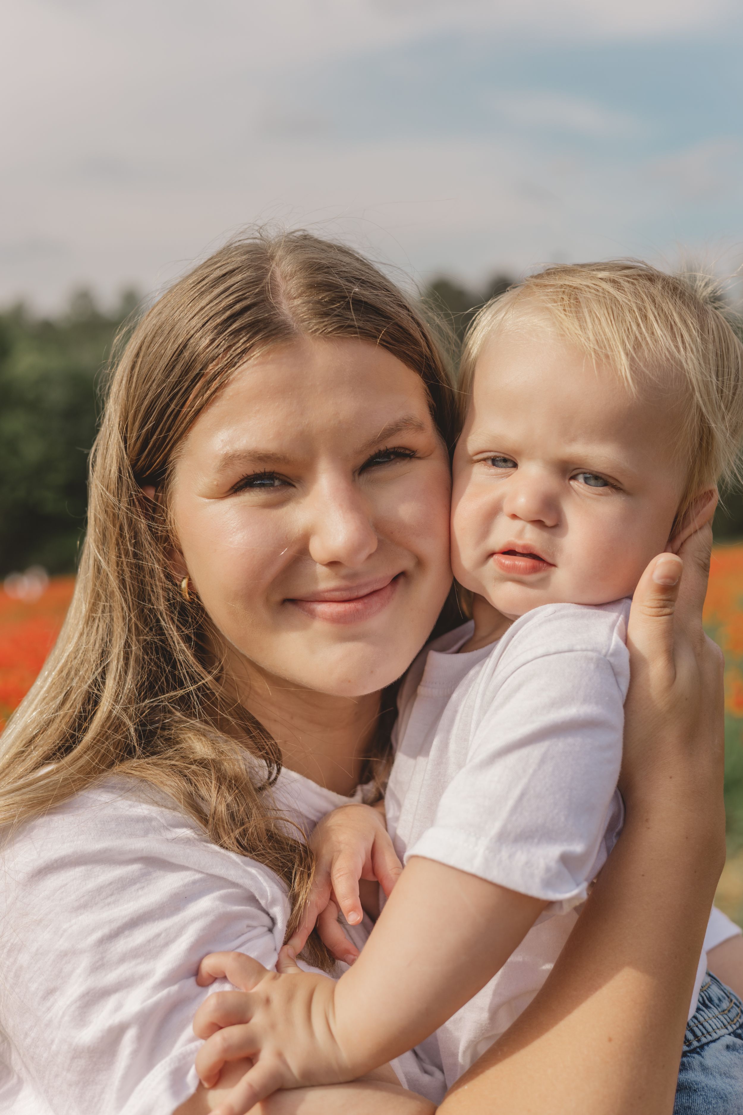 Smiling young woman in white shirt holding grumpy blonde toddler boy in poppy field on sunny day.