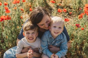 Mother hugs two young children while sitting in vibrant red poppy field outdoors.