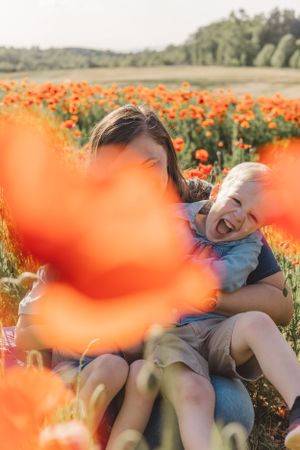 Mother and child laugh joyfully together sitting among bright red poppies in field.