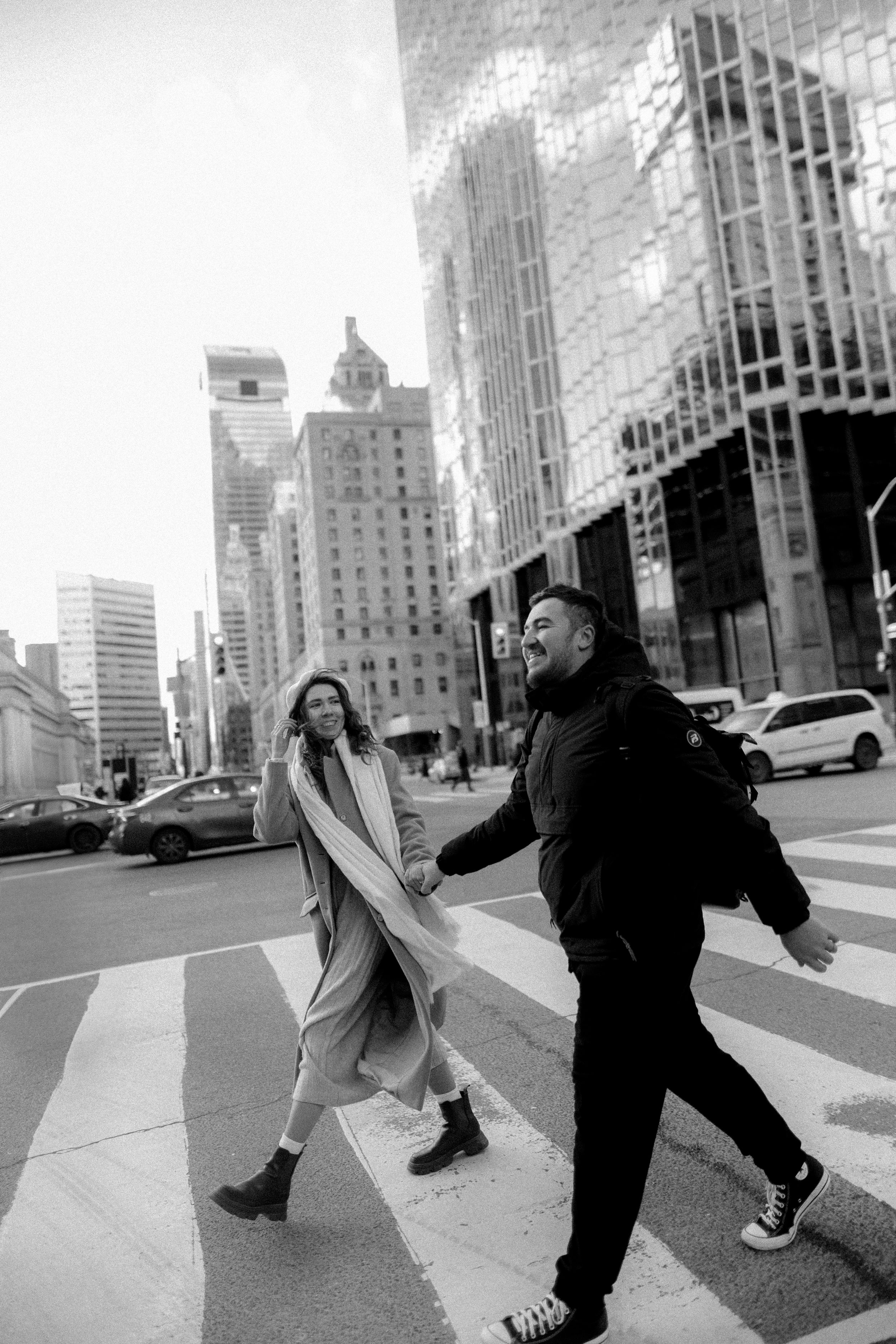 A couple crossing a city street with urban architecture in black and white.