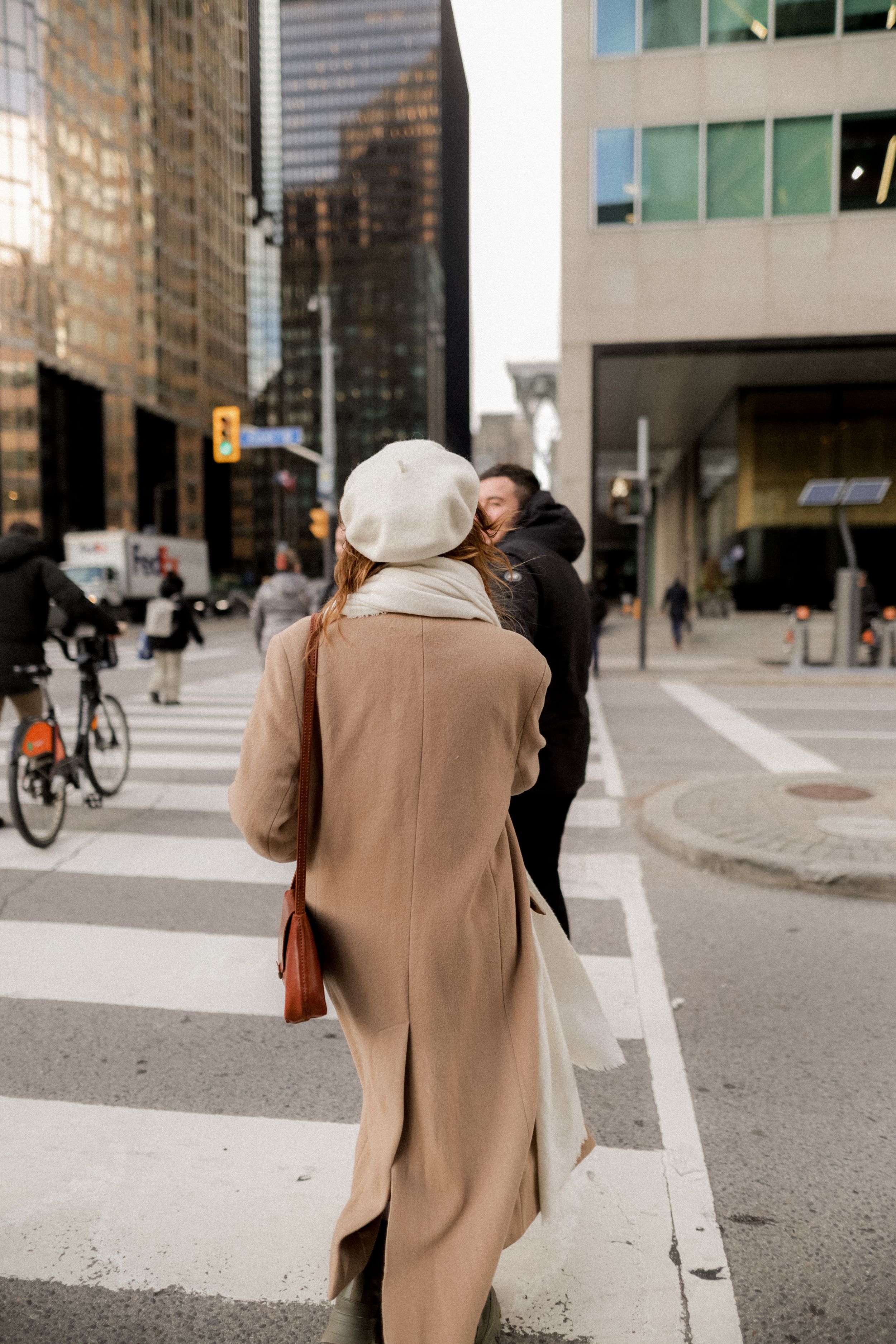 A person in a beige coat walking across a downtown crosswalk with office buildings towering above.