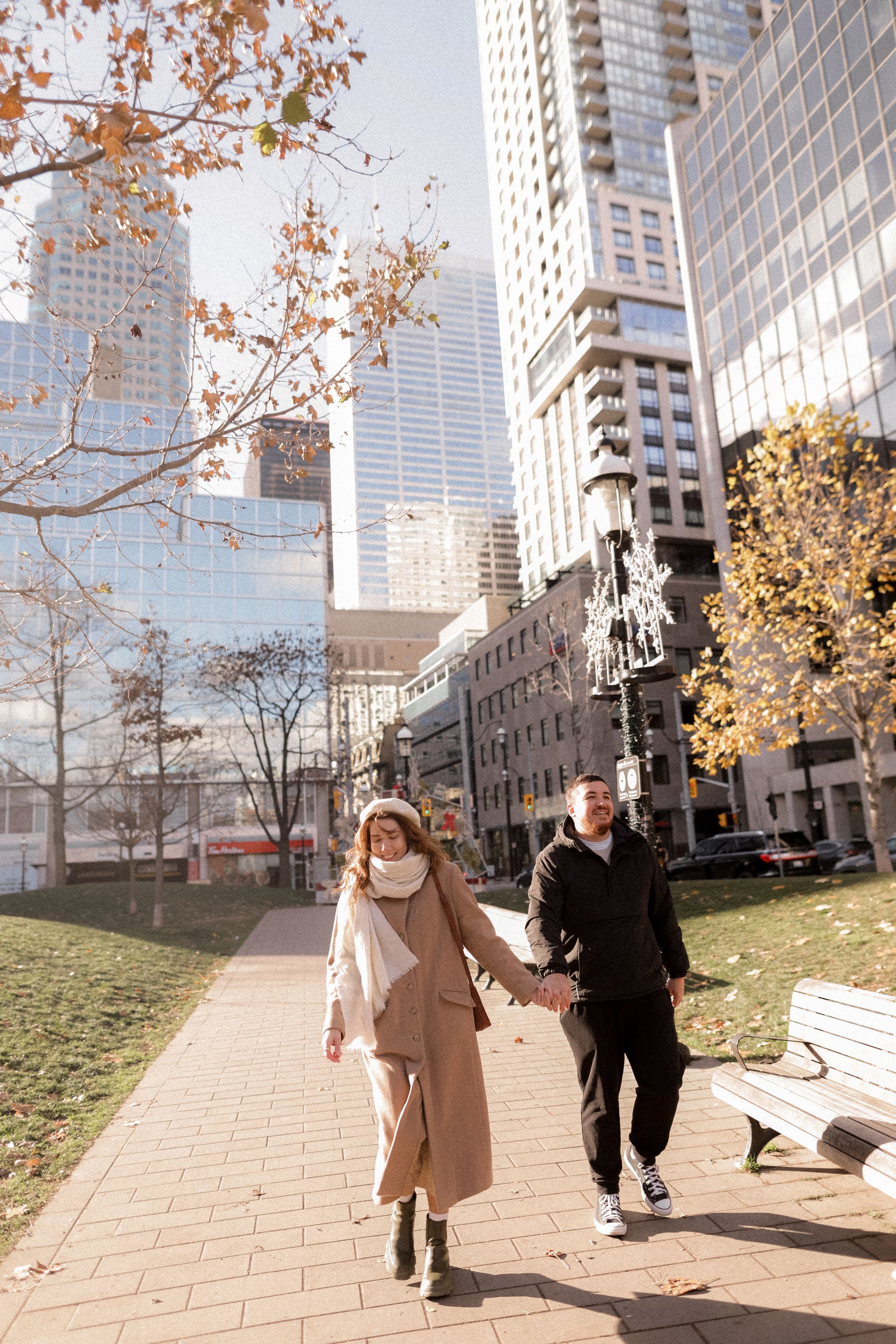 A couple walking hand in hand along a city path surrounded by autumn trees and skyscrapers.