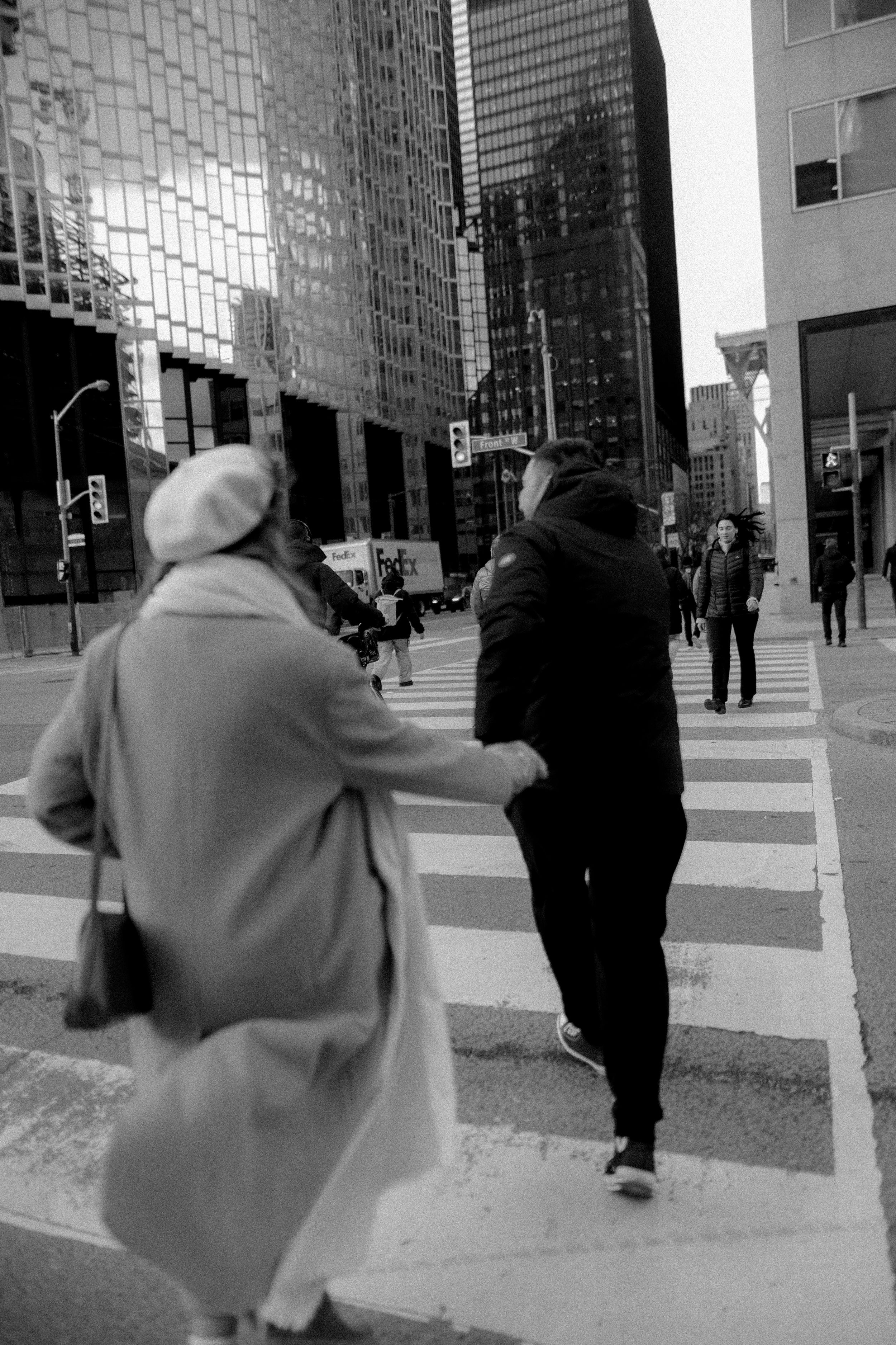 Two people in winter coats walking through an urban intersection with modern buildings in the background.