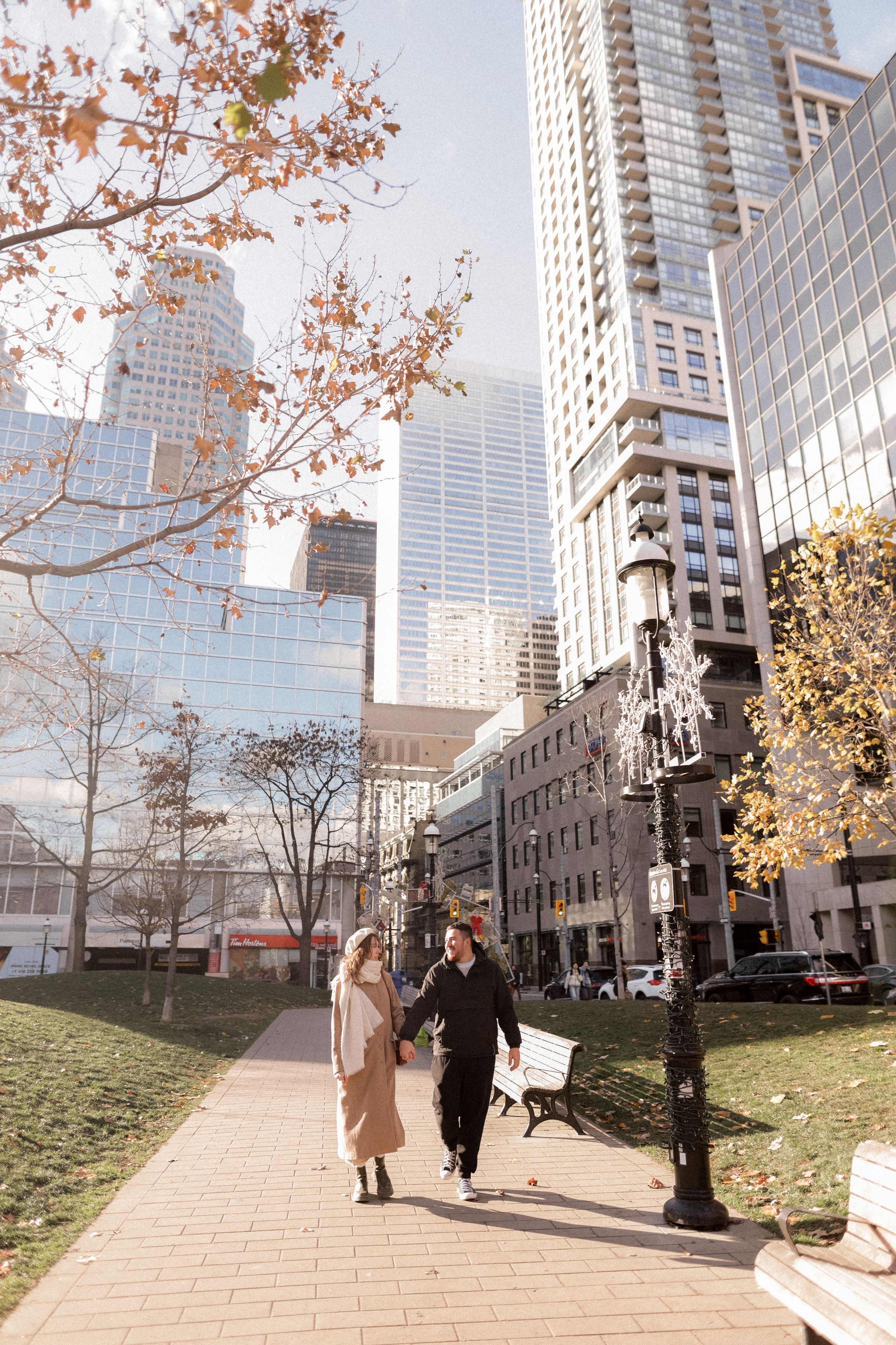 A couple walking along a downtown path with modern buildings and autumn trees.