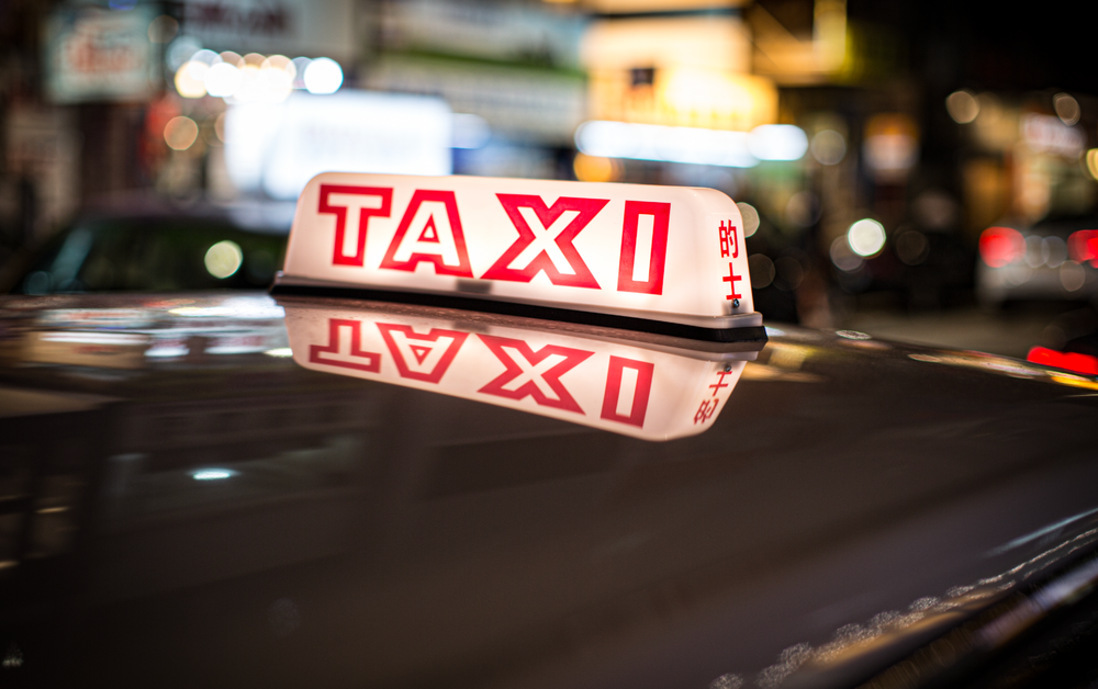 Illuminated red and white taxi sign on car roof glowing at night in urban street.