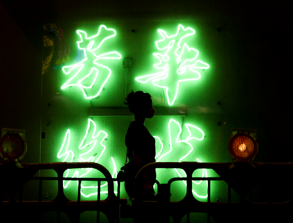 Silhouette of person standing before bright green glowing Chinese neon signs in darkness.