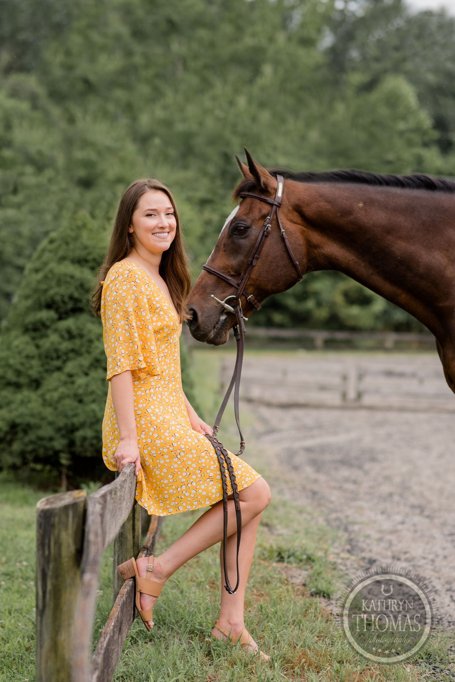 A brown horse and a person in a yellow floral dress pose together by a wooden fence in a rustic outdoor setting.