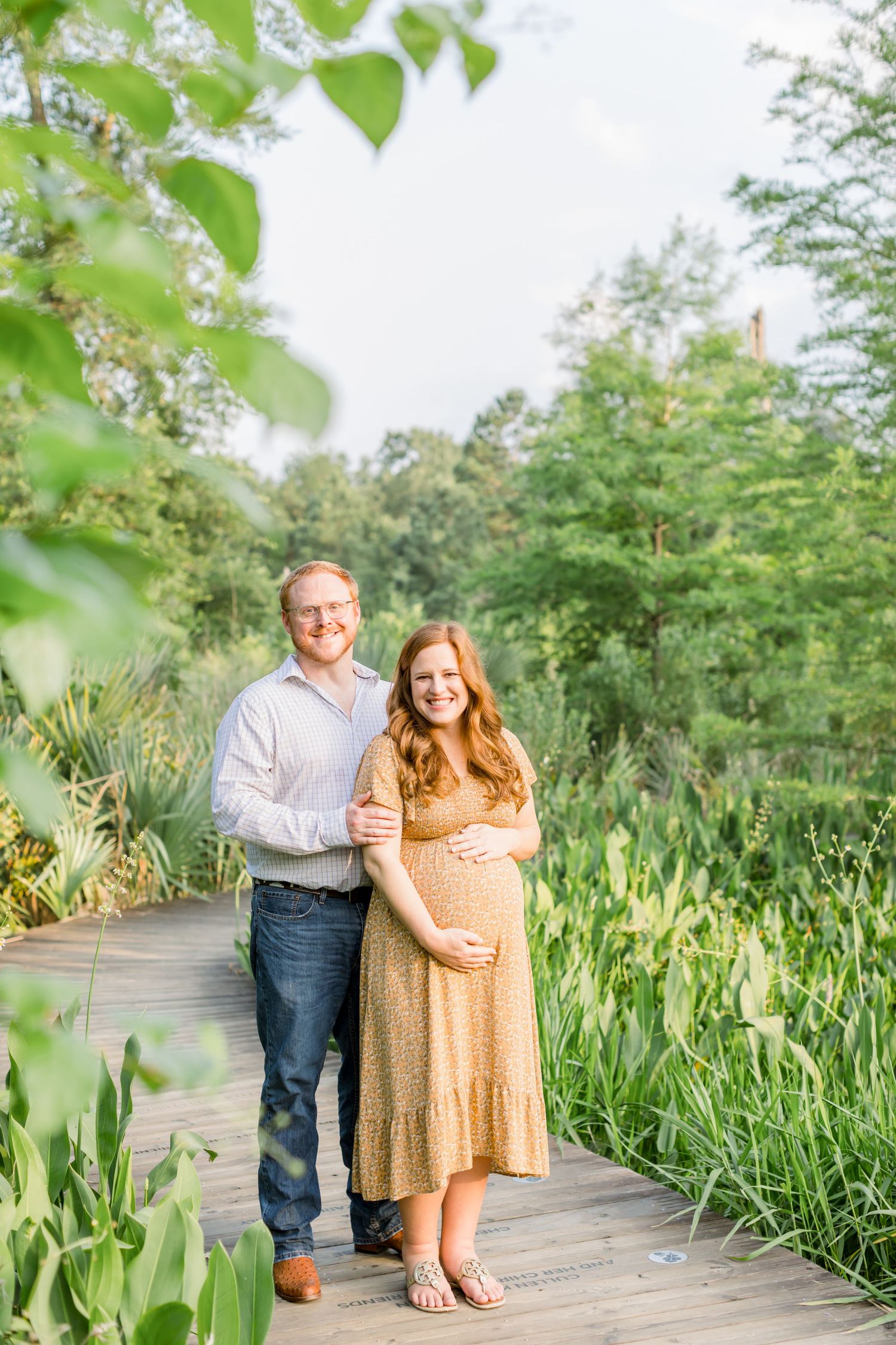 Maternity photo on wooden boardwalk with green foliage and trees in background.