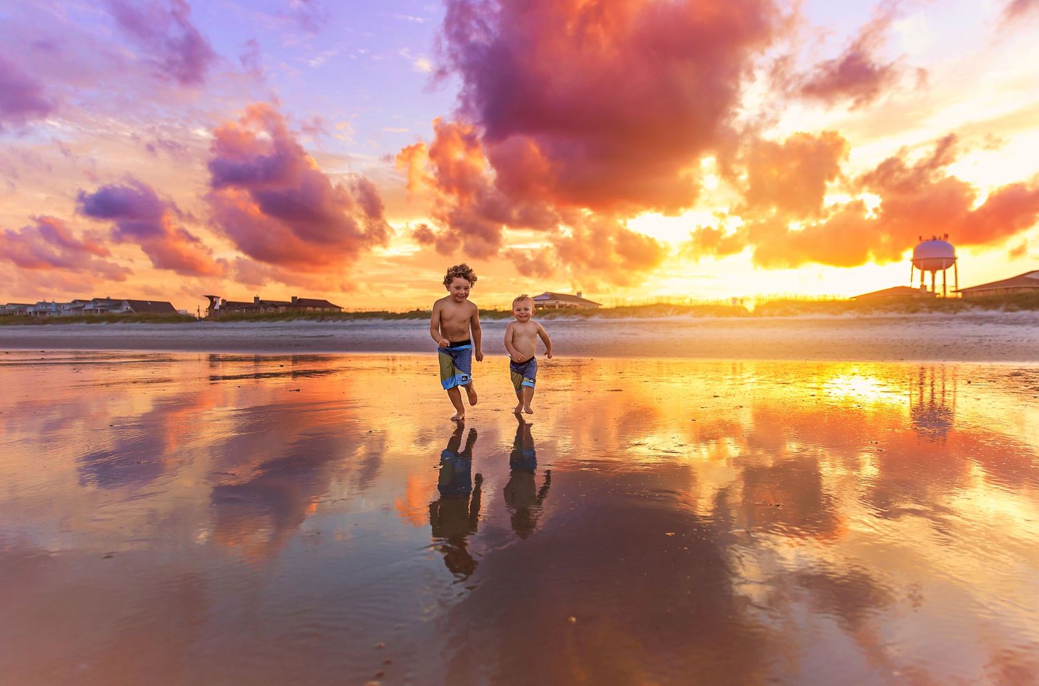 Two people walking on a beach at sunset with dramatic orange clouds reflected in wet sand.