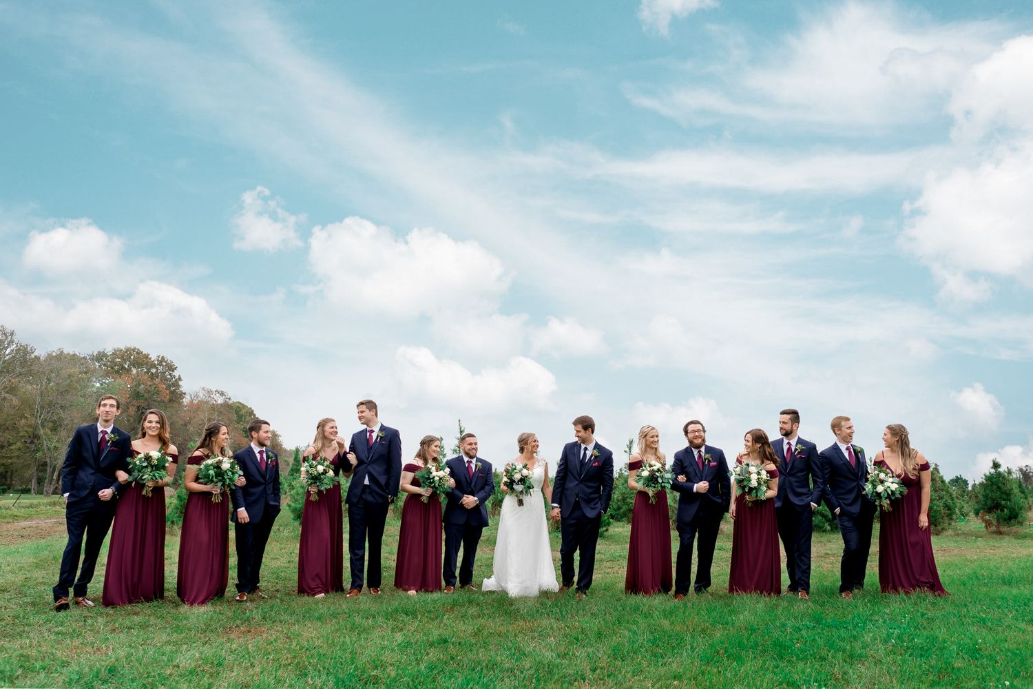 Wedding party in burgundy and navy poses on green field under bright blue sky.