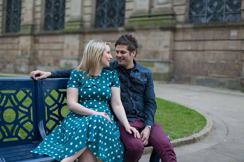 A man and woman sit on a park bench in front of a stone building, looking lovingly at each other.