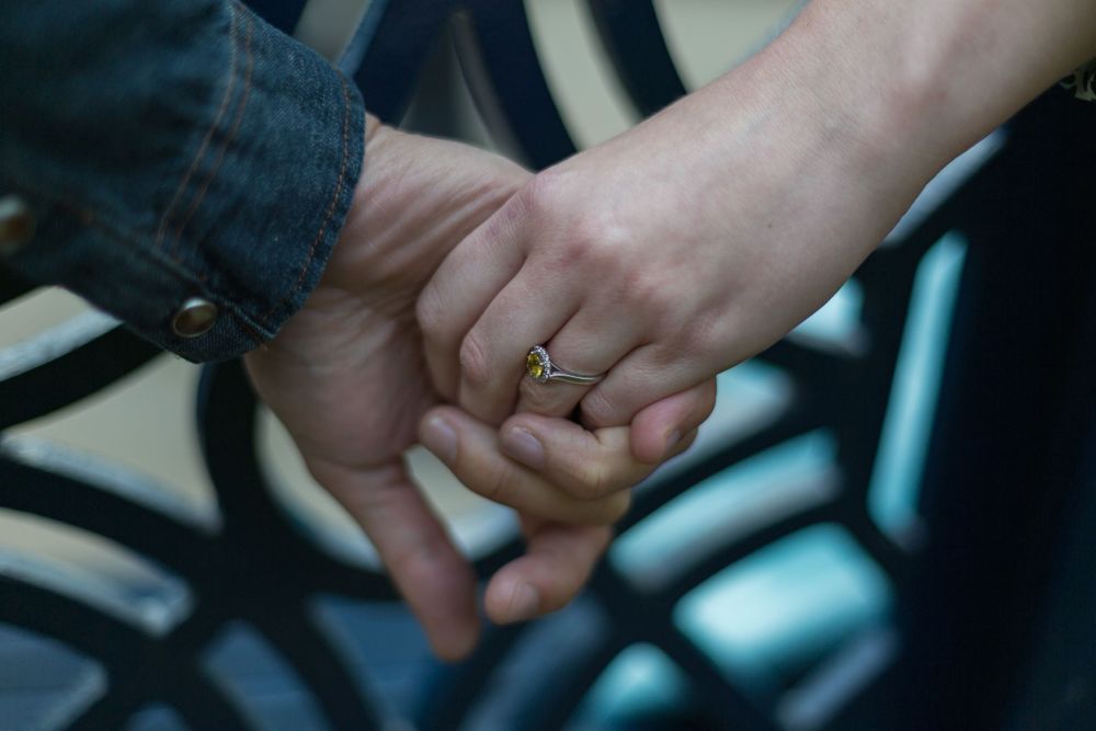 A close-up of a man and a woman holding hands behind a park bench. An engagement ring visible on the woman's hand.