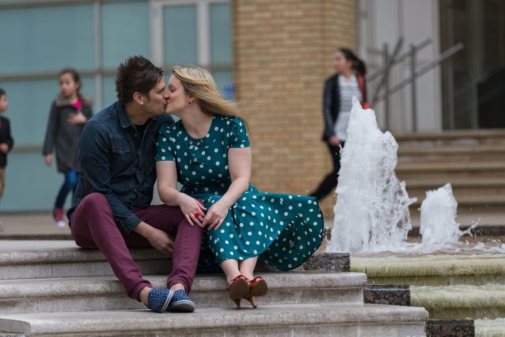 A man in a blue shirt and burgundy trousers and a woman in green and white polkadot dress kiss, next to a fountain.