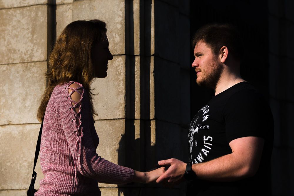 A woman in a pink sweater holds hands with a man in a black t-shirt. Left side in light, right side in shadow.