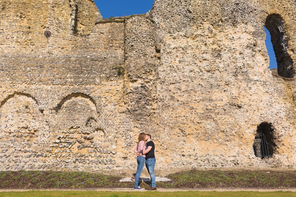 A man and a woman embrace and share a kiss in front of large stone ruins on a sunny day.