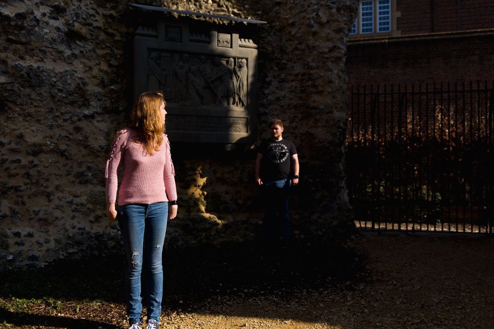 A woman in a pink sweater and blue jeans looks back towards a man, both highlighted by the sun amongst the shadows.