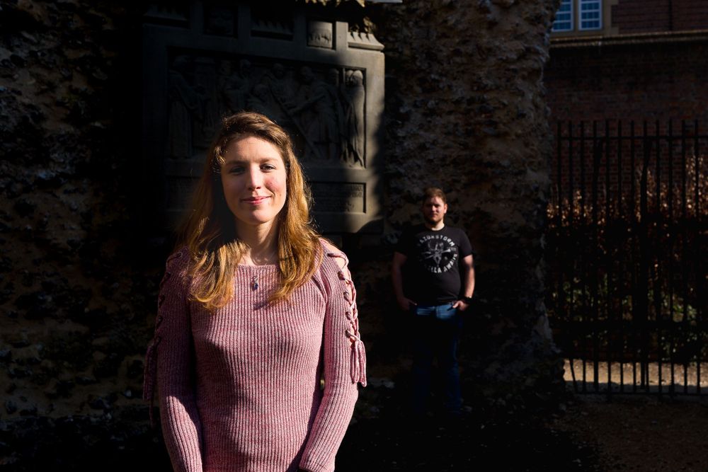 A woman in a pink sweater stands illuminated against a dark background. A man stands in a pocket of light behind her.
