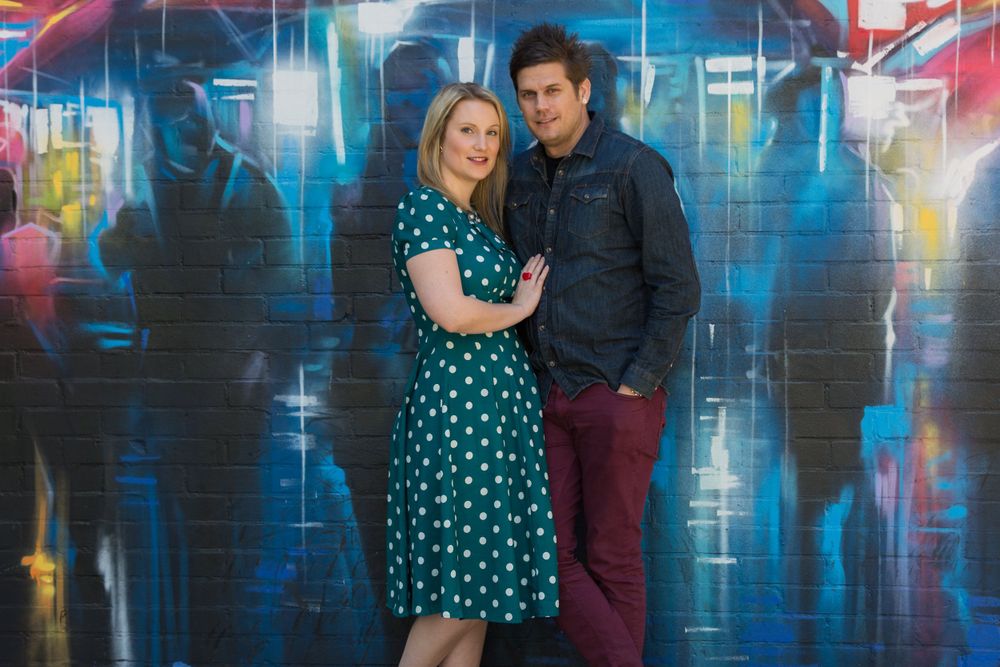 A newly engaged man and woman pose for a photo in front of some street art.