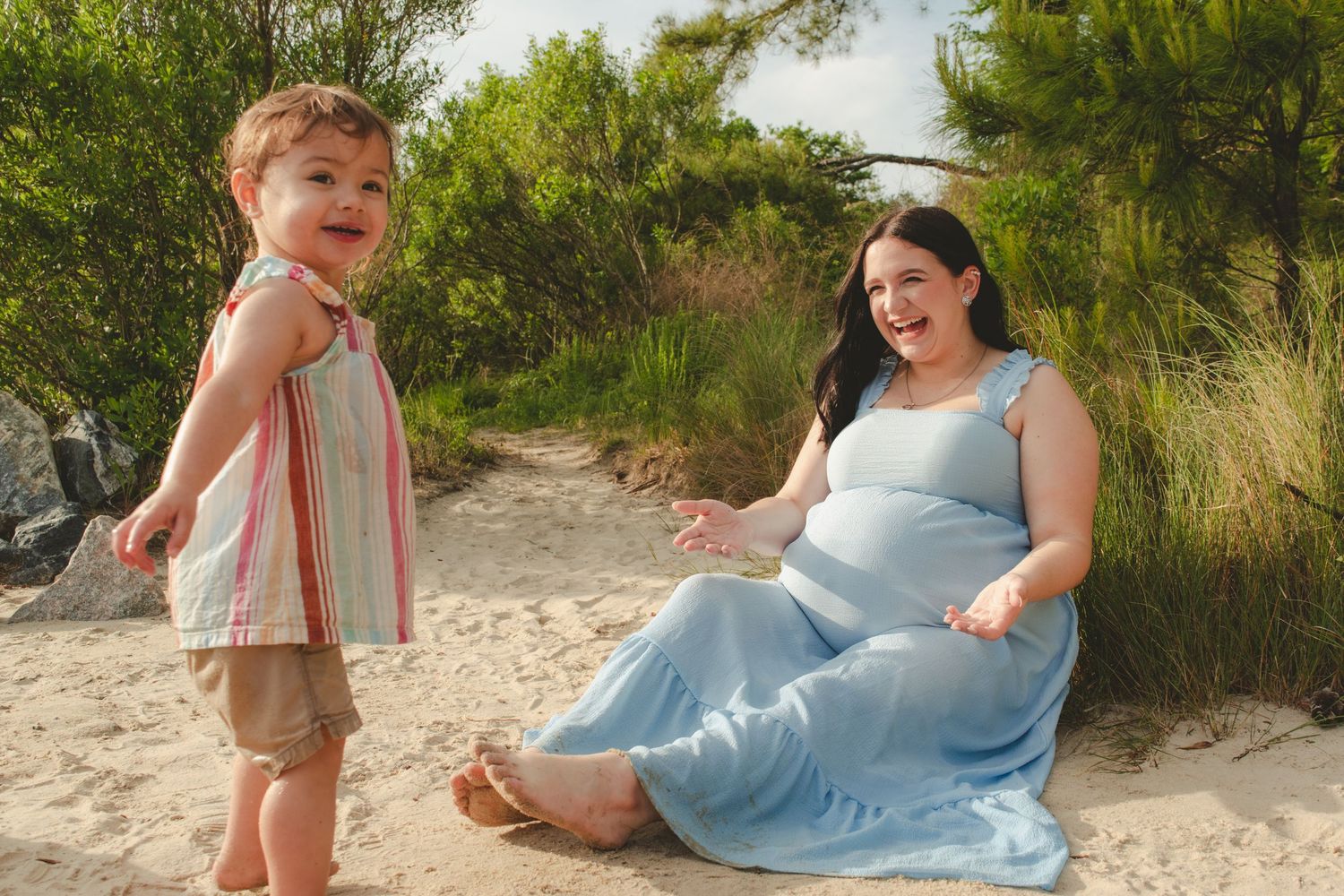 A small child stands on beach next to person in blue dress sitting on sand.