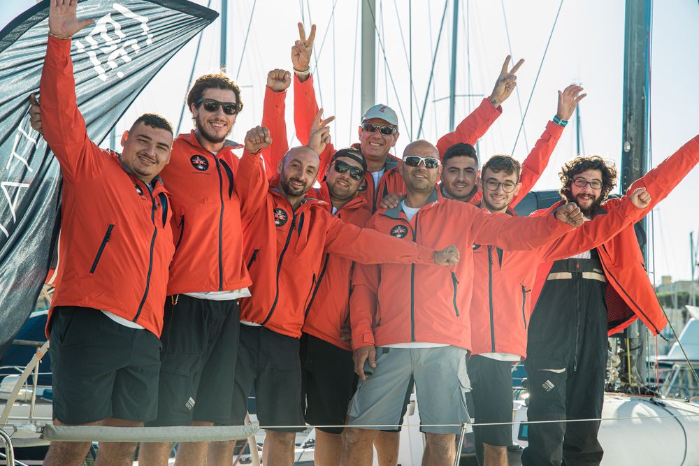 Celebrating sailing team in matching red jackets cheering with raised arms on a boat at the marina.