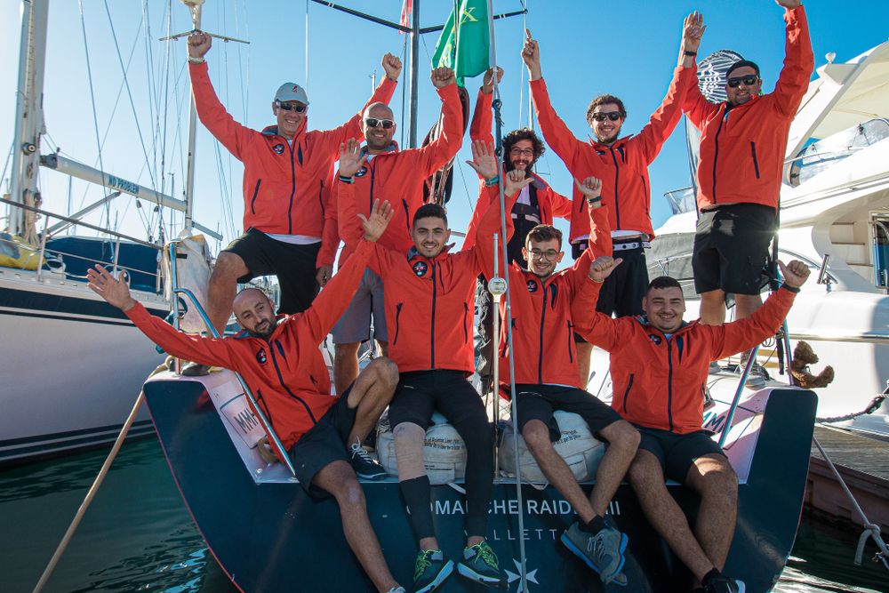 Victorious sailing crew in red uniforms posing joyfully on yacht deck with green flag at the marina.