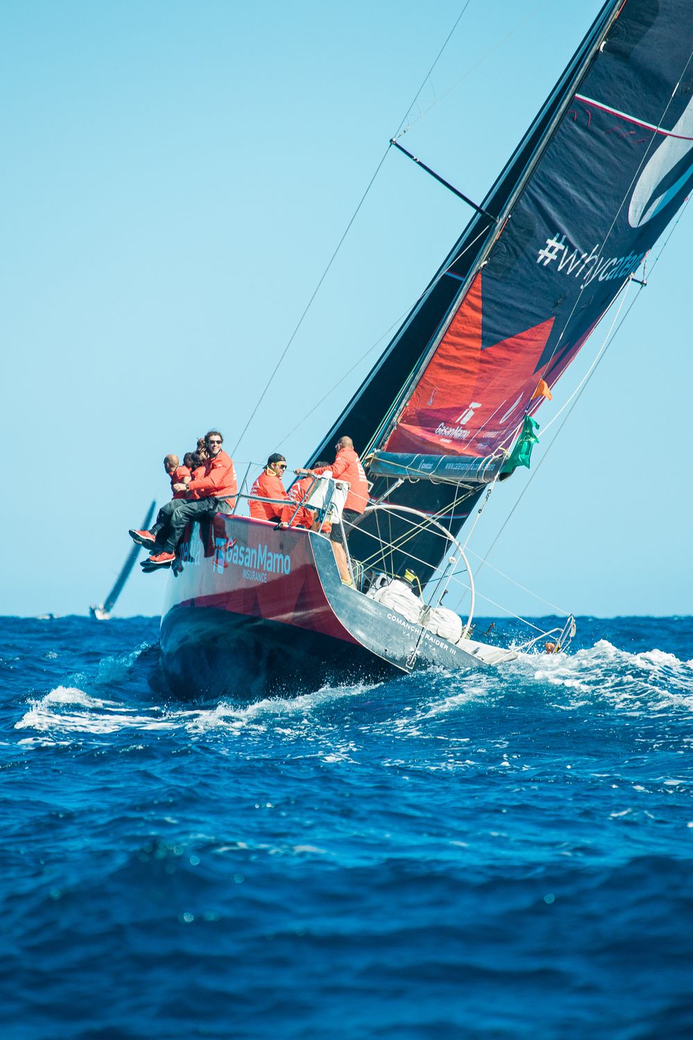 GasanMamo sponsored racing sailboat heeling sharply with crew hiking out in open sea.