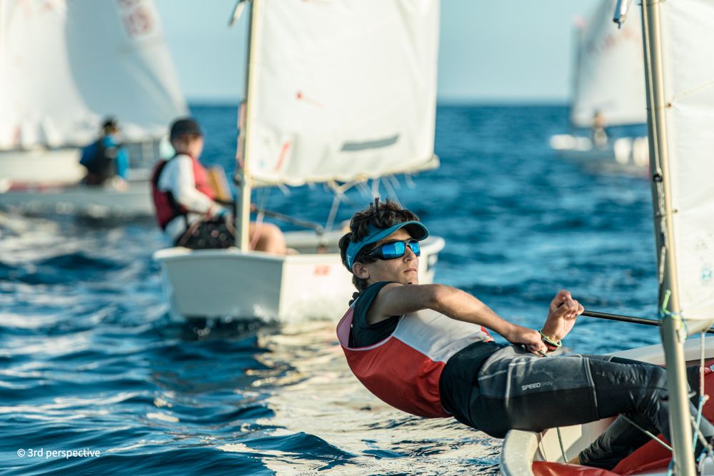 Young sailor leaning back on small Optimist dinghy wearing goggles during sunny regatta race.