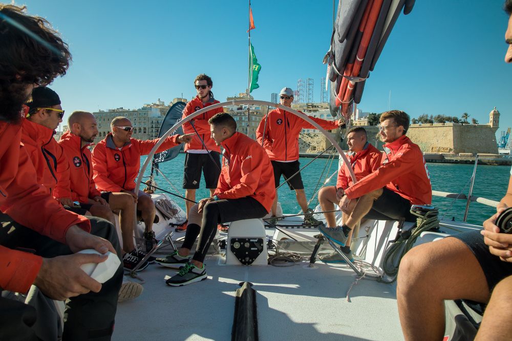 Sailing team in red gear gathered on yacht deck discussing race strategy in a sunny Mediterranean port.