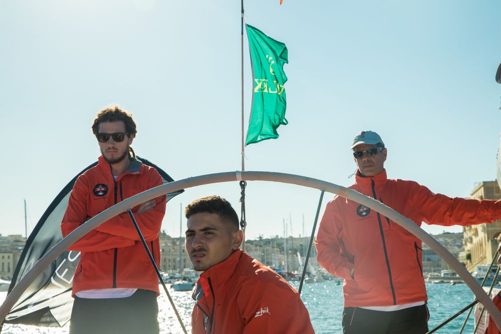 Three sailors in red jackets navigating a sailboat in a sunny harbor with historic waterfront backdrop.