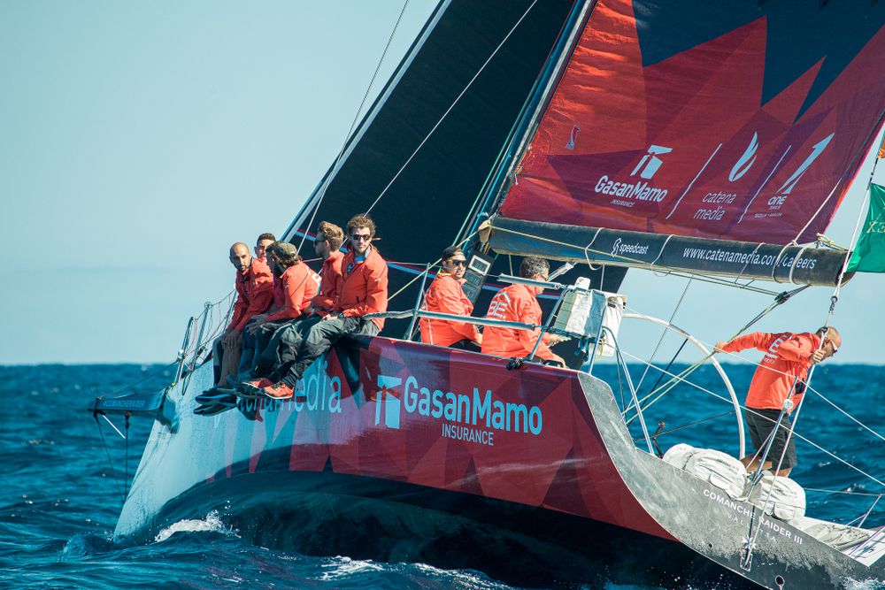 Red GasanMamo sailing yacht racing through blue ocean waters with crew in orange life jackets.
