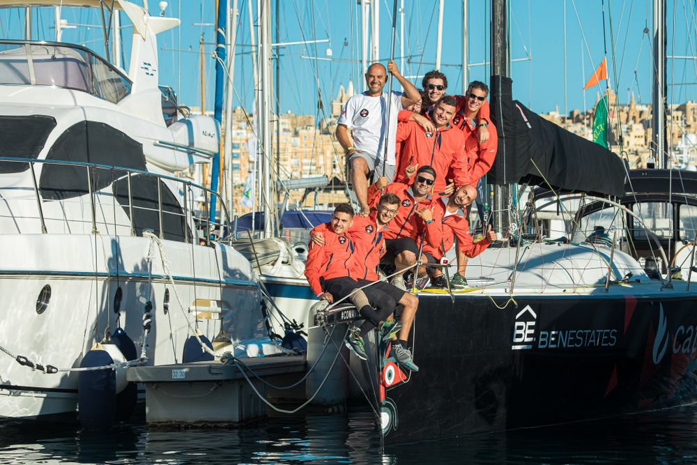 Sailing team in red uniforms posing on dark racing yacht docked in sunny Mediterranean harbor.
