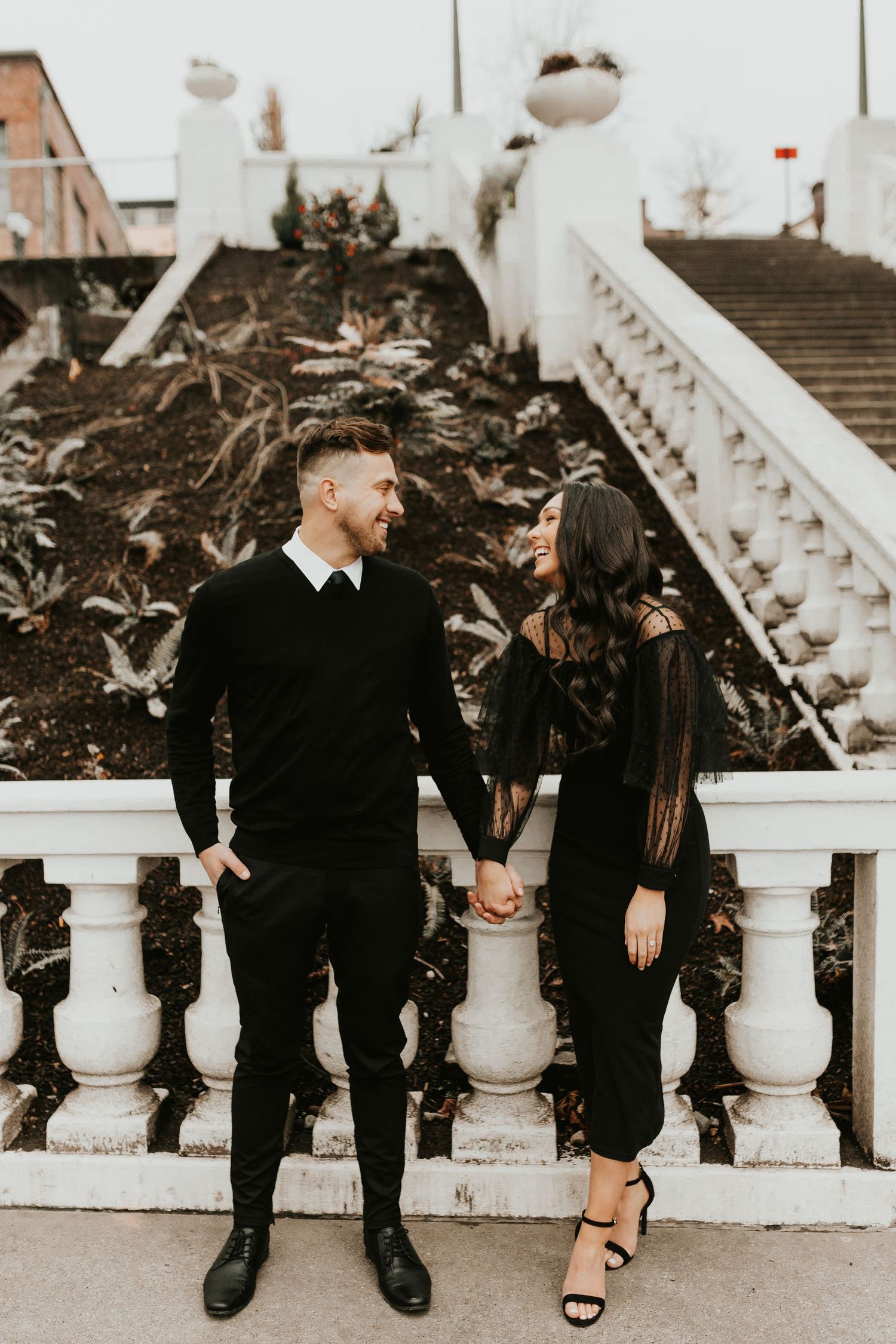 Stylish couple in black outfits poses on white stone staircase in vintage-toned photo.