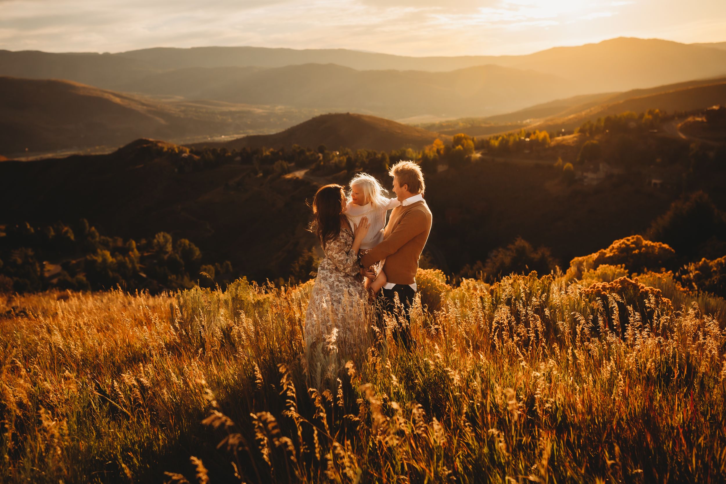 A family of three candidly smiles together at golden hour with a Vail mountain vista behind them