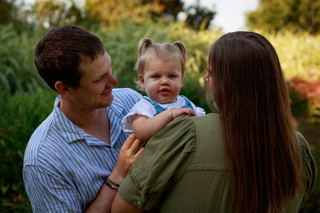 A family of three stands close together outdoors, surrounded by lush greenery, sharing a warm and joyful moment.